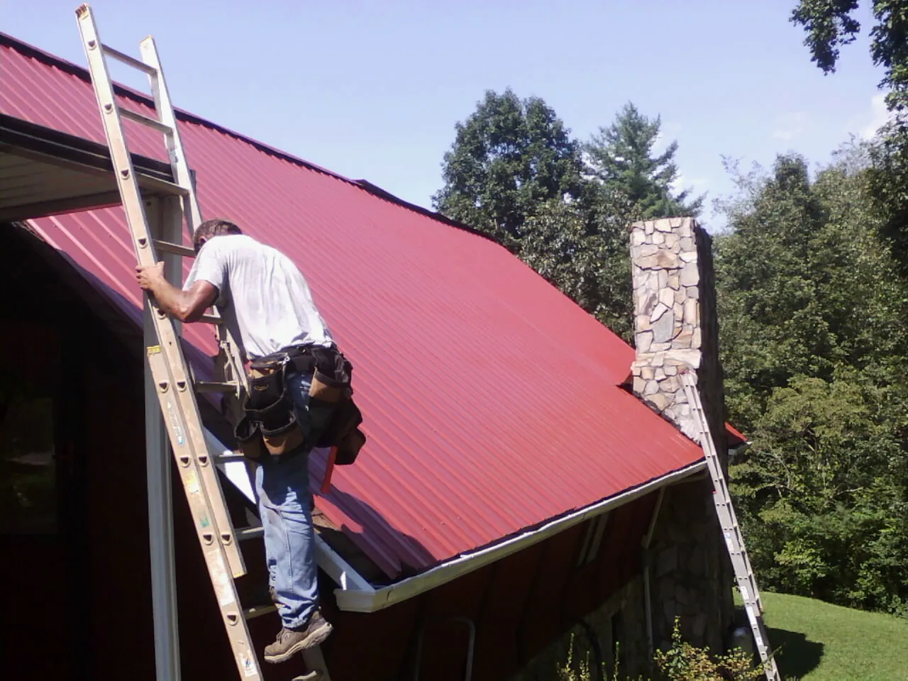 Roofer on a ladder beside a red metal roof and stone chimney — Soffit & Fascia Repair by Roofing Brothers in Amsterdam